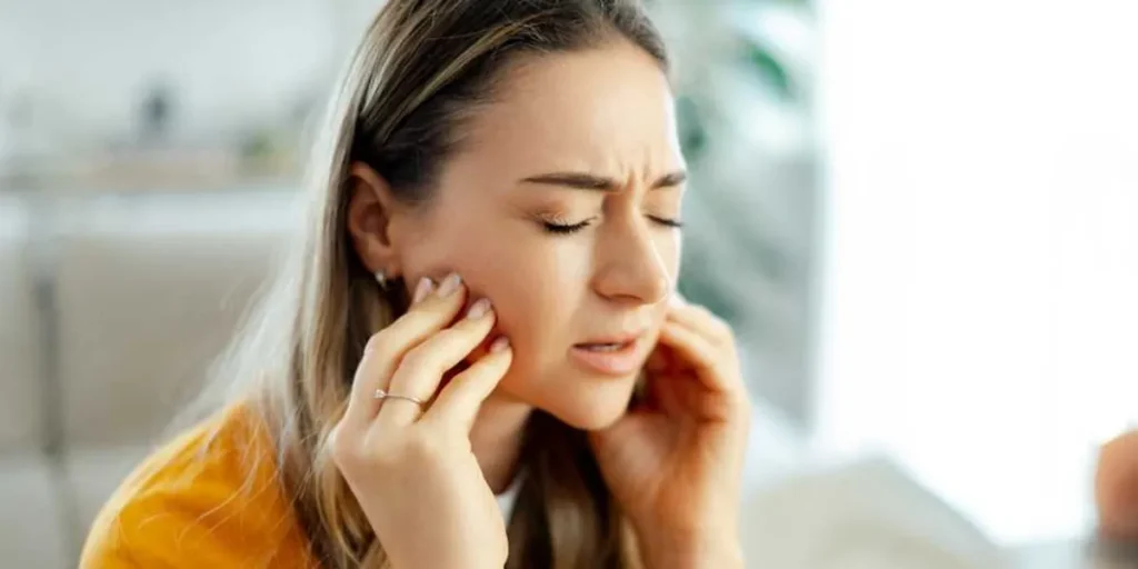 woman in yellow shirt experiencing jaw pain eyes closed hands on cheeks